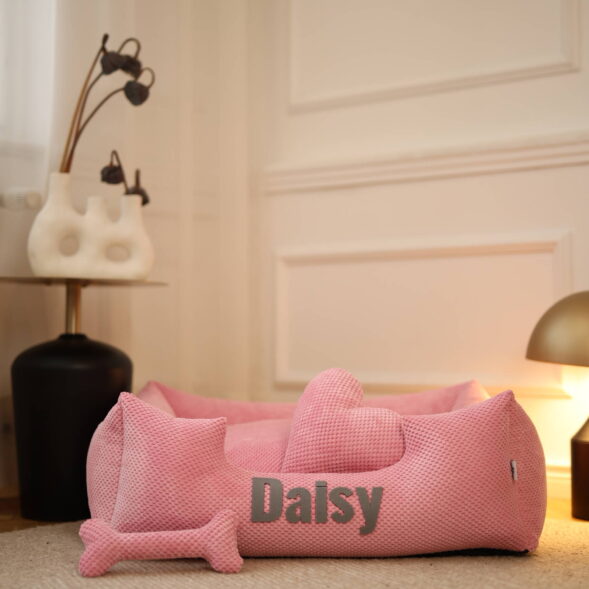 Różowe legowisko Daisy – personalizowany komfort dla Twojej suczki Różowe legowisko Daisy z poduszką w kształcie kości, ustawione w stylowym salonie
